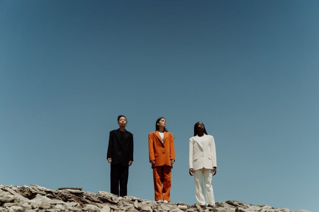 Three people in colored suits stand on rocky terrain against a clear blue sky, showcasing modern fashion.
